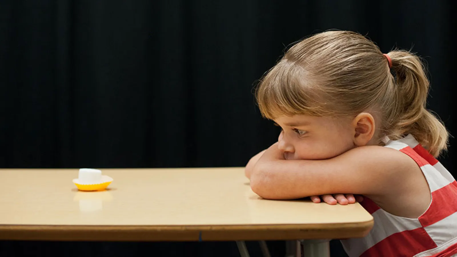 Little girl staring at marshmallow across the table from her with arms crossed.