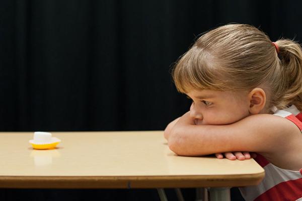 Little girl staring at marshmallow across the table from her with arms crossed.